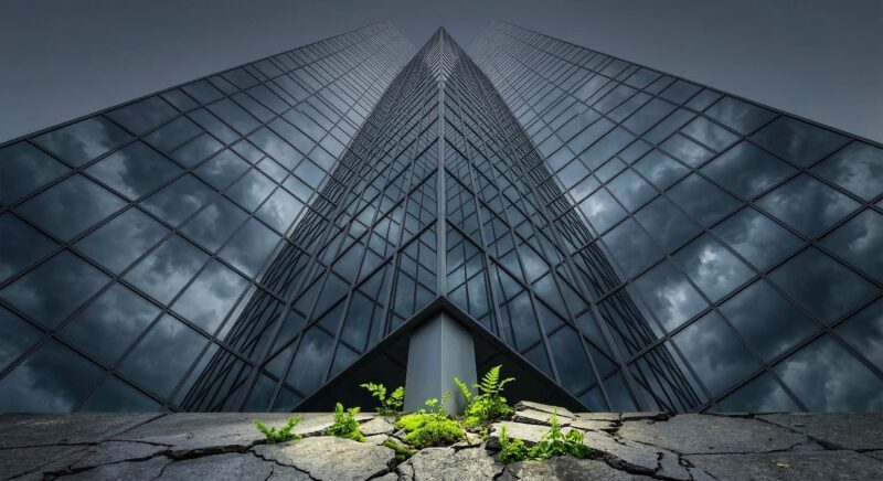 a towering glass corporate skyscraper seen from below, its reflective surface showing storm clouds. At the base of the building, the sidewalk is cracked and green plants are pushing through the concrete 